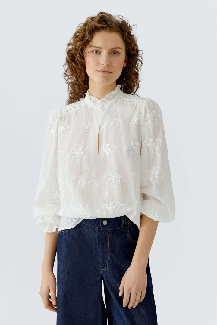 Femme portant une Blouse à broderies florales blanche avec col montant et manches bouffantes.