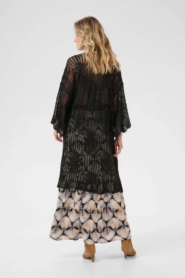 Cardigan Franca long en tricot ajouré noir, fluide, porté sur une jupe à motifs floraux.