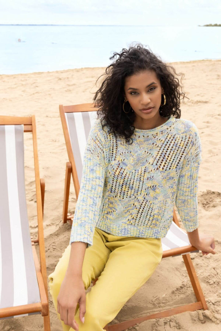 Femme portant un chandail ajouré à motif texturé, assise sur une chaise longue à la plage, avec un look décontracté et soigné.