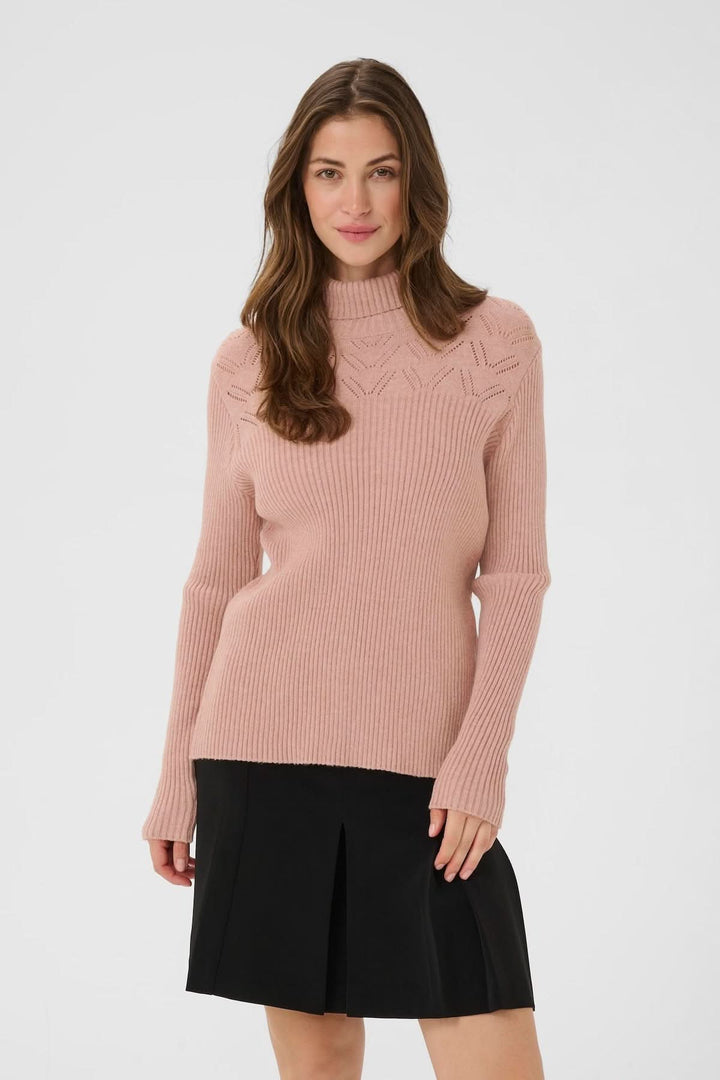 Femme portant un chandail à col montant en maille côtelée rose avec détails ajourés, combiné avec une jupe noire élégante.
