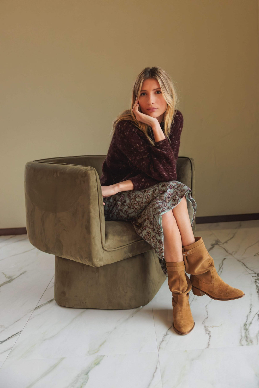 Femme assise portant un chandail maille texturée marron avec jupe imprimée et bottes camel dans un fauteuil moderne.