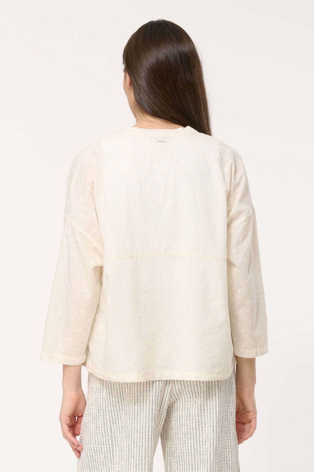 Femme portant une chemise légère en coton brodé, vue de dos, avec un pantalon rayé, élégante et décontractée.