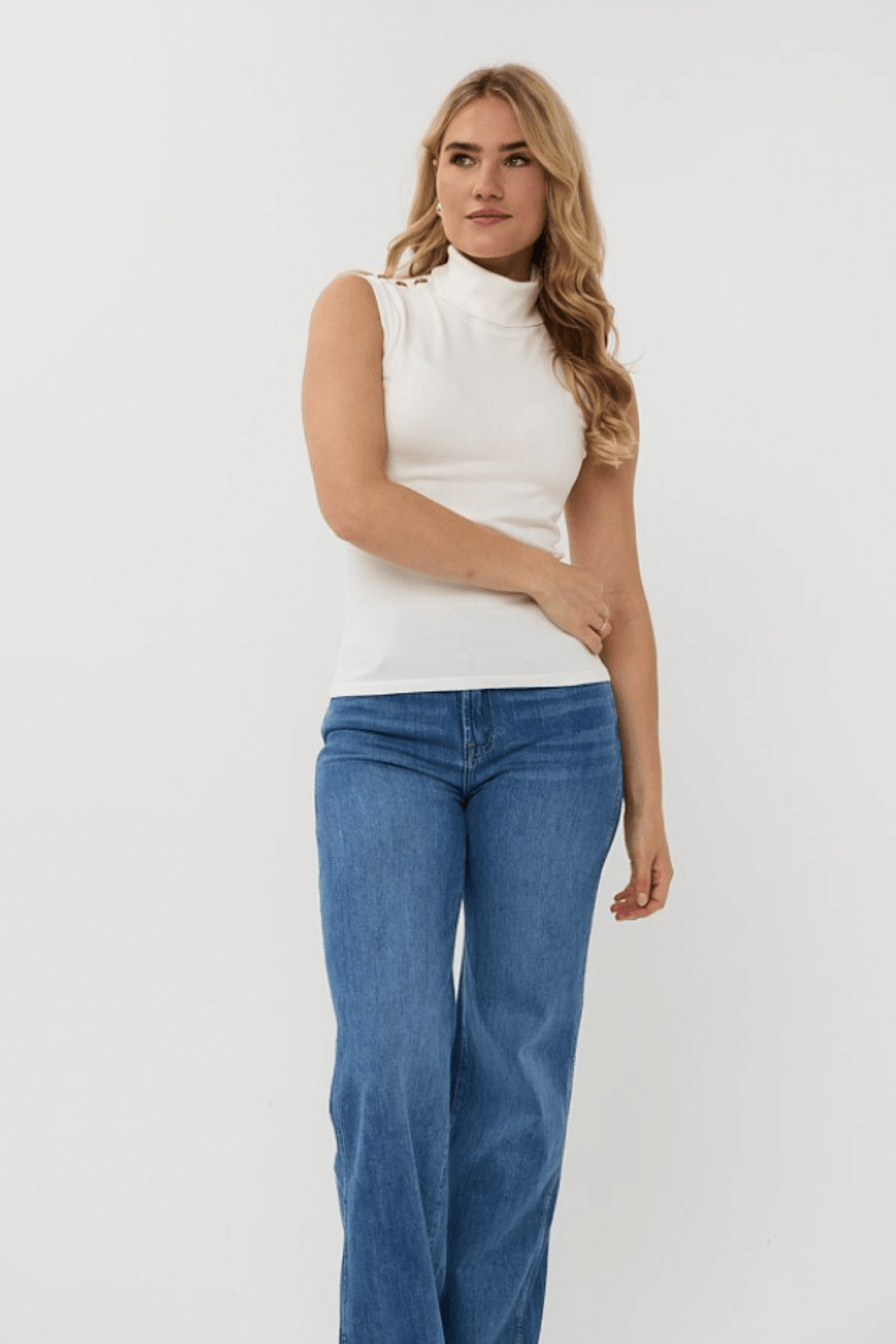 Col roulé sans manches blanc avec boutons métalliques, porté avec un jean bleu pour un look chic et élégant.