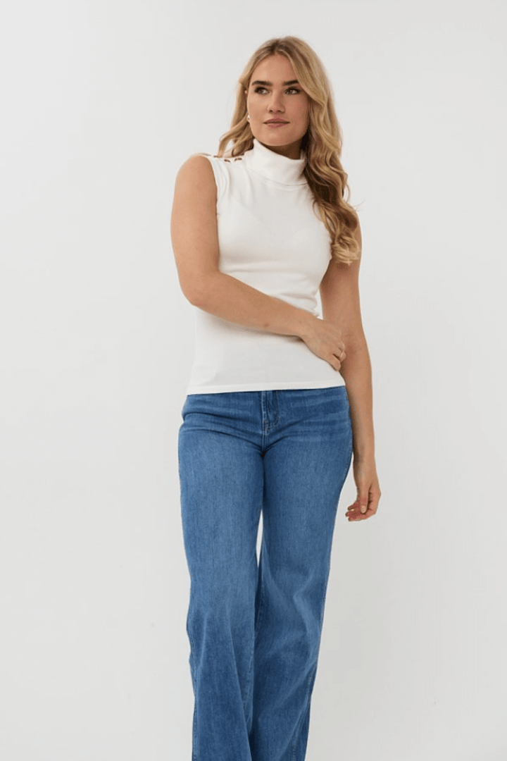 Col roulé sans manches blanc avec boutons métalliques, porté avec un jean bleu pour un look chic et élégant.