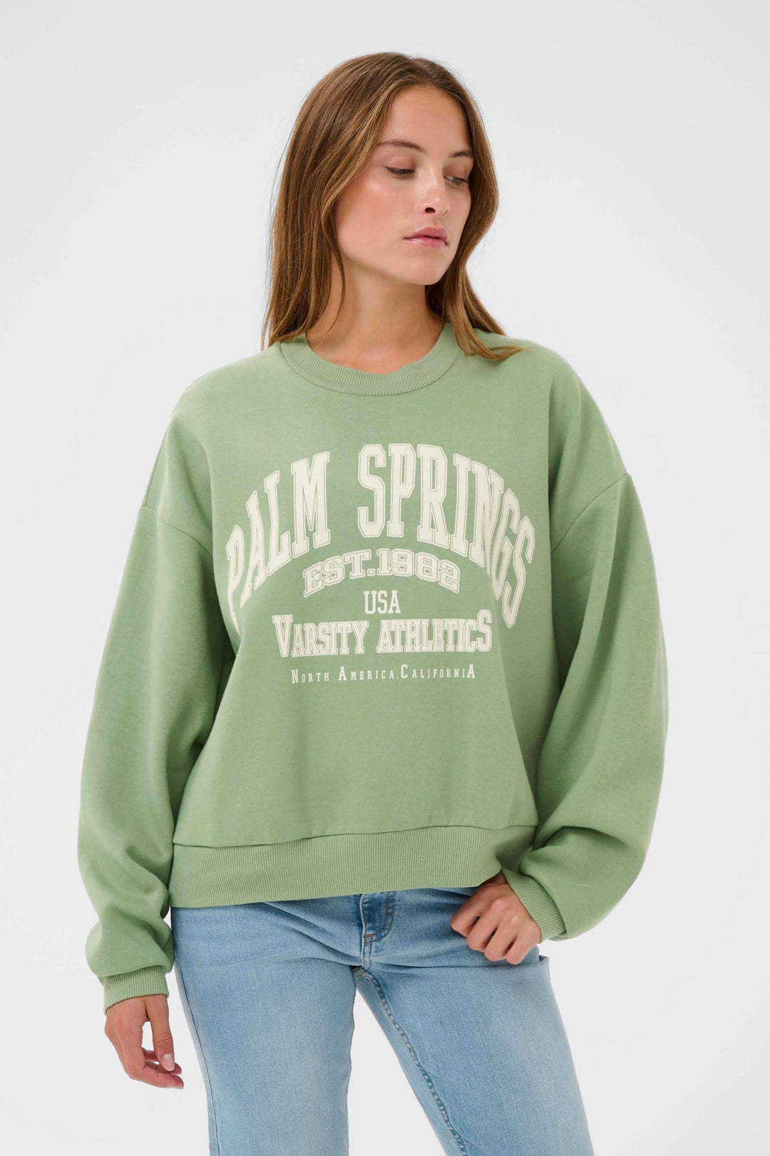 Jeune femme portant un coton ouaté Sussi vert avec motif universitaire, coupe ample et confortable, style décontracté et tendance.