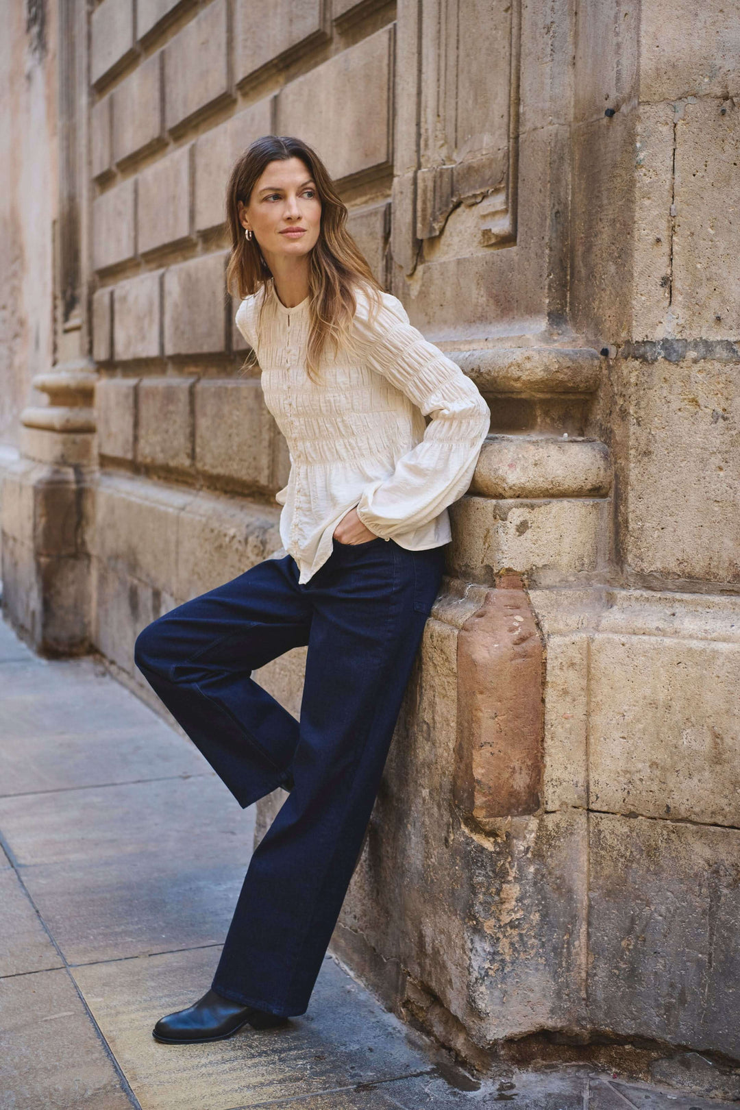 Modèle portant le Jean Simona taille mi-haute avec une chemise blanche, posant contre un mur dans une rue pavée.