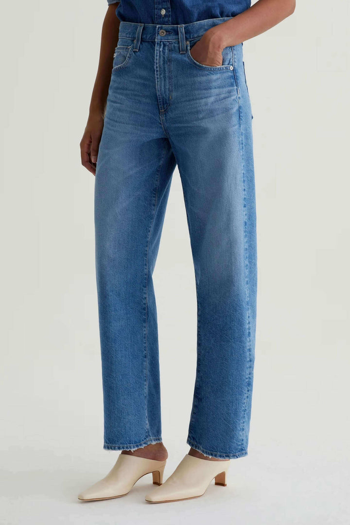 Jean droit à jambe large en denim bleu avec taille haute, décontracté et moderne, porté avec des talons blancs pour un style minimaliste.