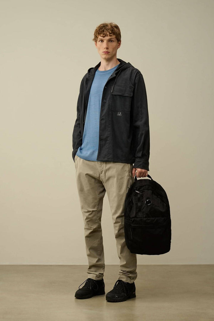 Homme portant un pantalon beige et un hoodie noir, tenant un sac à dos noir, dans un environnement neutre.