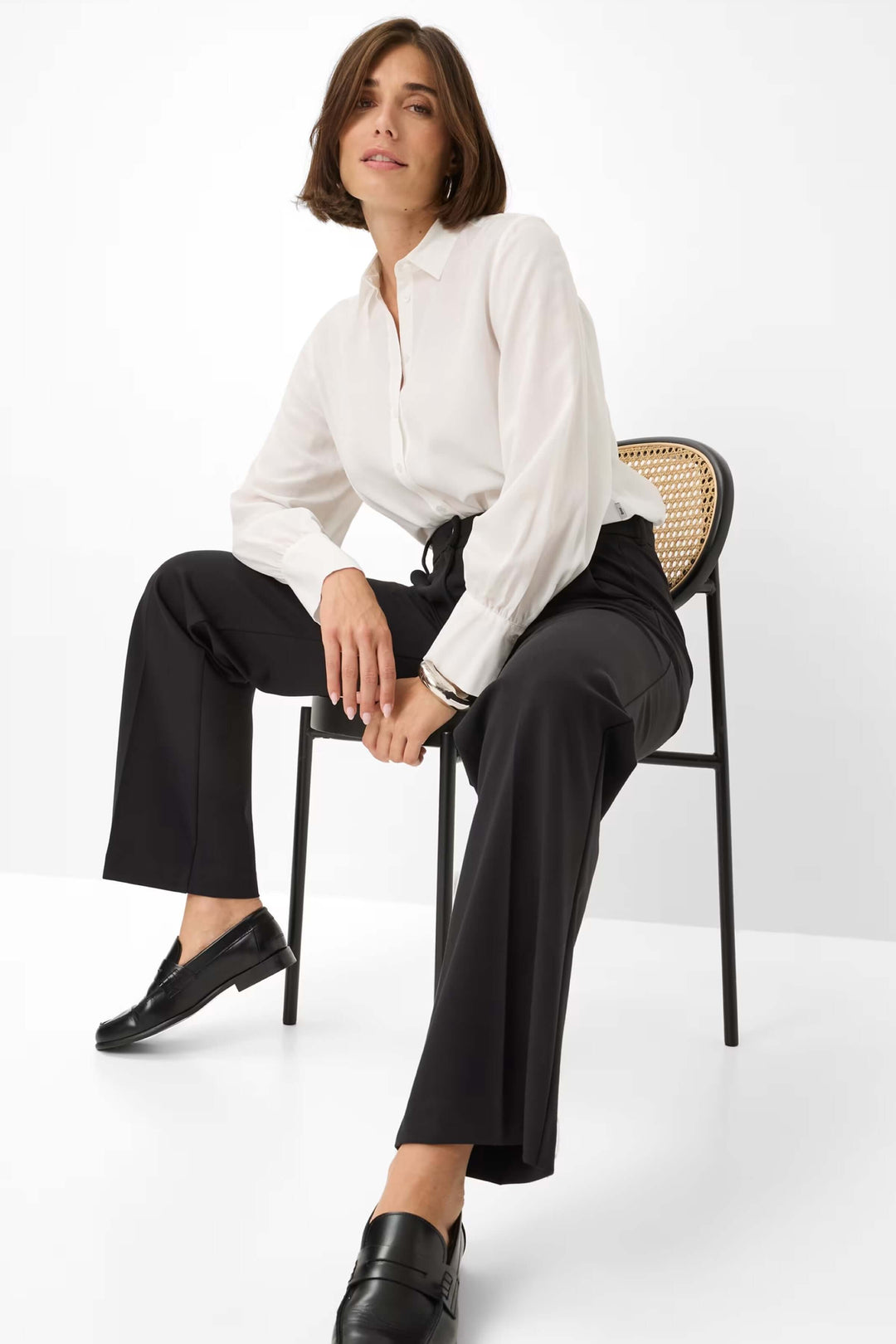 Femme portant un pantalon palazzo noir avec une chemise blanche, assise sur une chaise. Mode élégante et confortable.