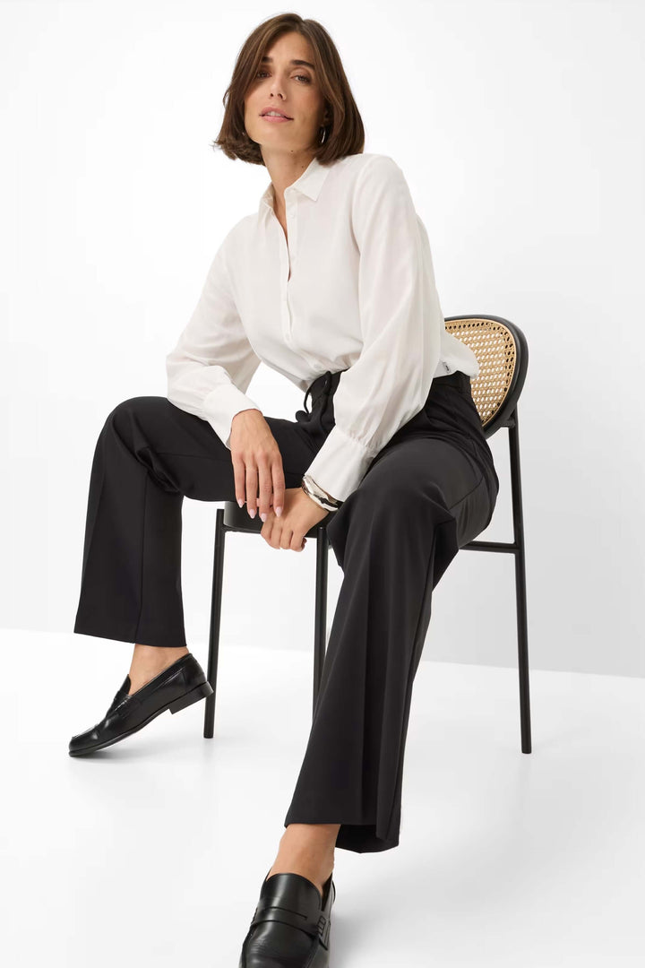 Femme portant un pantalon palazzo noir avec une chemise blanche, assise sur une chaise. Mode élégante et confortable.