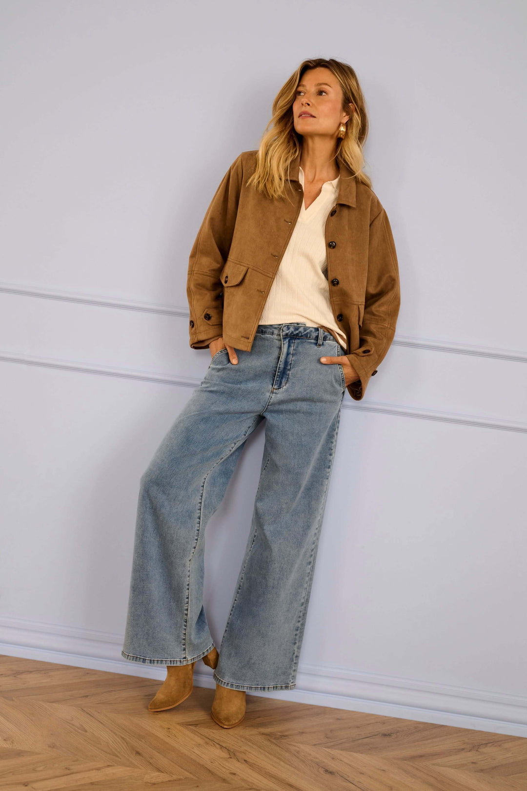 Woman wearing wide-leg jeans, beige jacket, and suede boots leaning against a light blue wall.