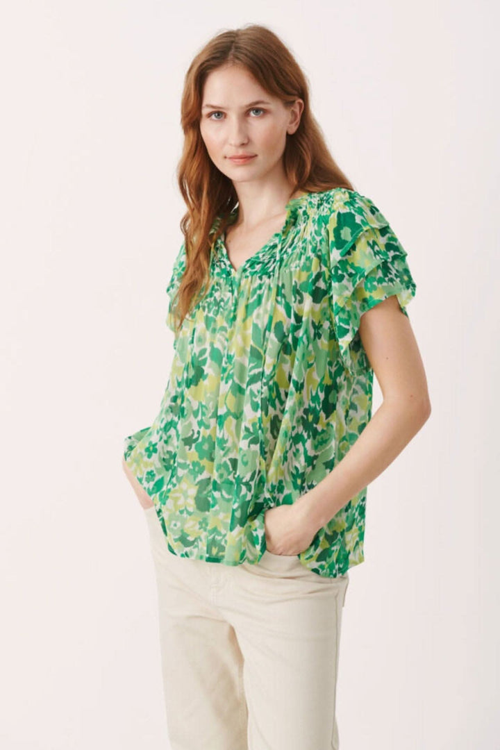 Femme portant une blouse fleurie Sinna en vert avec des manches courtes et des petits volants.