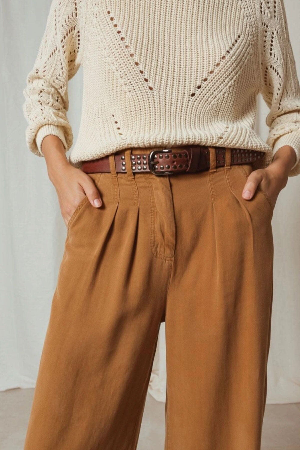 Ceinture en cuir clouté marron portée avec un pull crème et pantalon camel, ajoutant une touche moderne à la tenue.