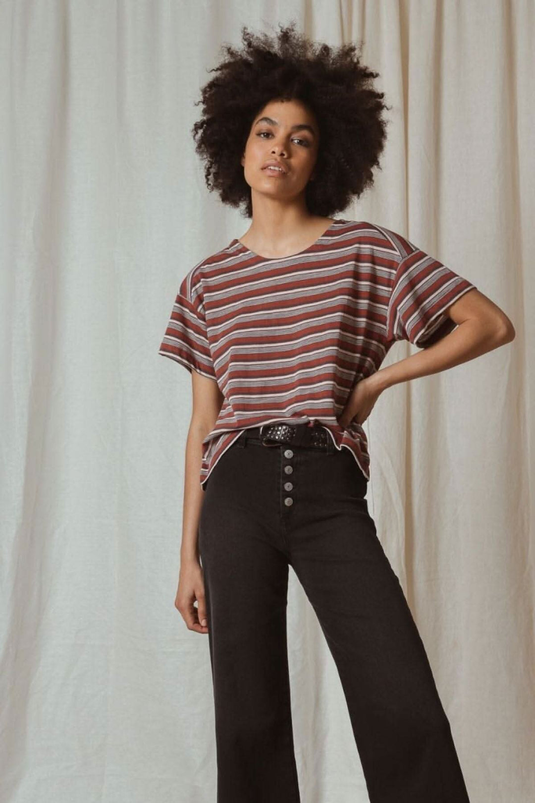 Femme stylée avec cheveux crépus, portant un t-shirt rayé et un pantalon noir devant un fond beige.