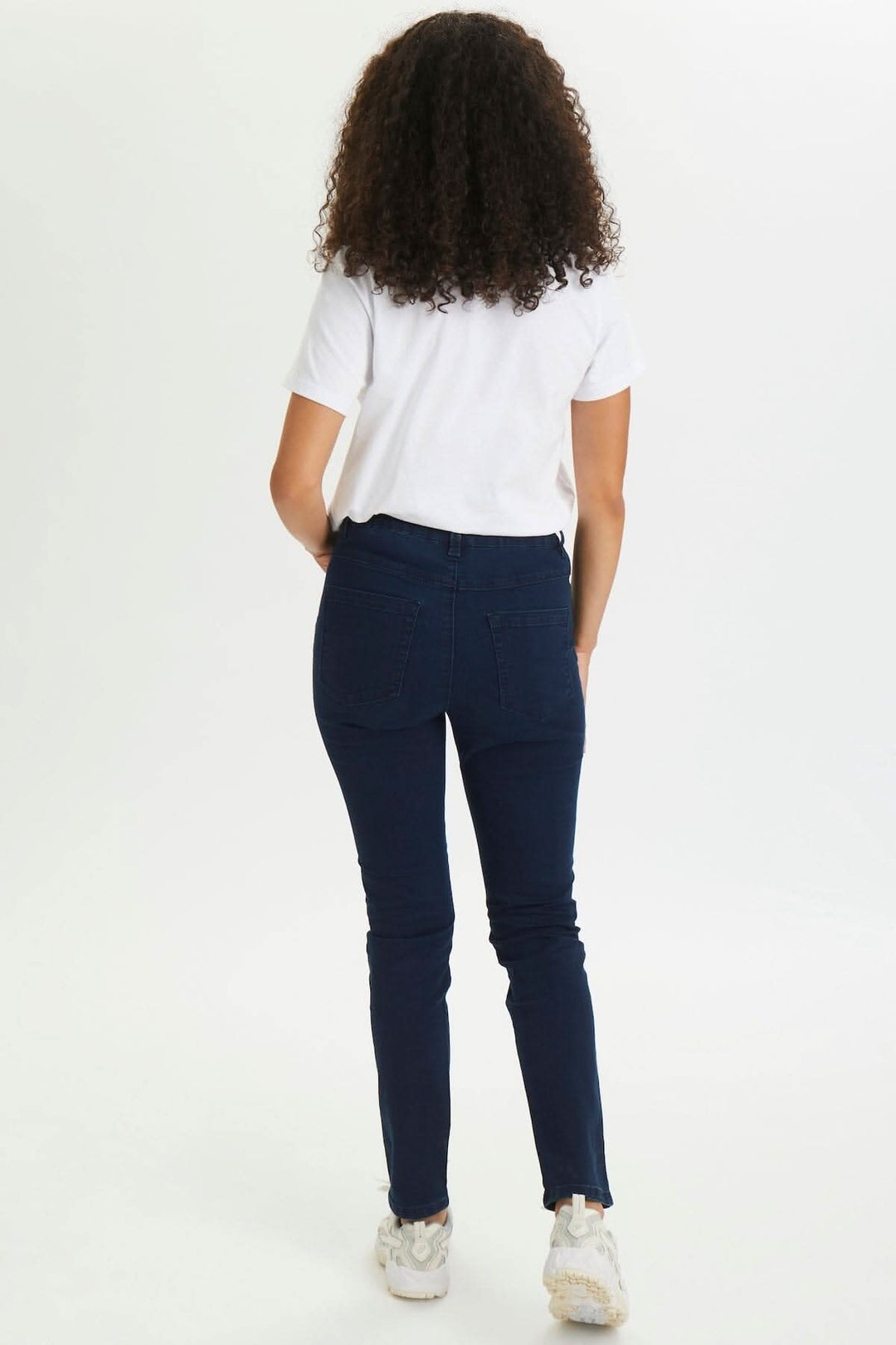 Femme portant un jean étroit de couleur foncée, vue de dos, avec un t-shirt blanc et des chaussures blanches.