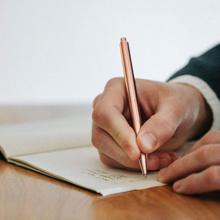 Main image of a hand writing with a pink crayon Modern Fuel on a blank notebook.