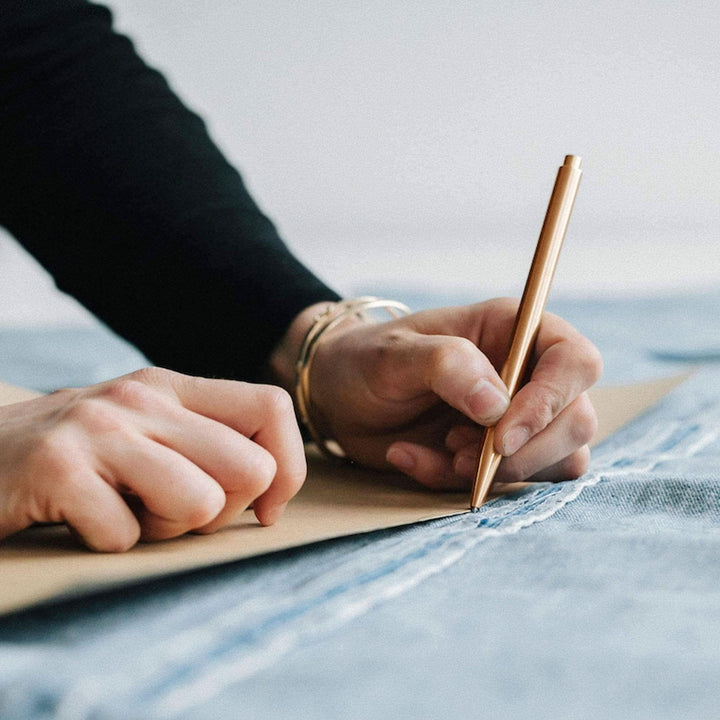 Personne utilisant un crayon Modern Fuel doré pour dessiner sur du papier, avec un tissu en denim en arrière-plan.