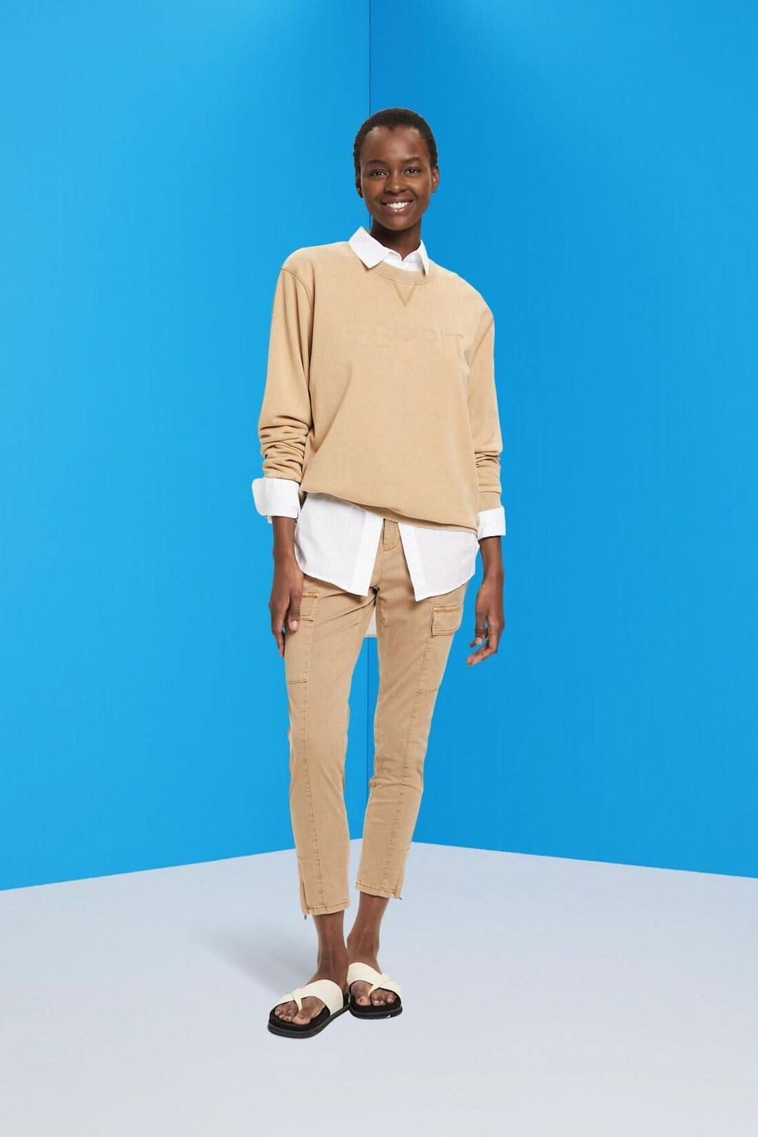 Modèle portant un pull beige sur une chemise blanche et un pantalon cargo, devant un fond bleu.