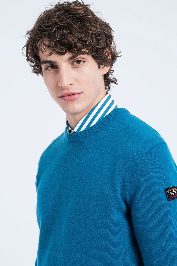 Pull en laine Shetland bleu, col rond, porté par un mannequin avec une chemise à rayures.